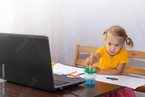 An interested little girl looks at her laptop and drawing picture. Online lessons.