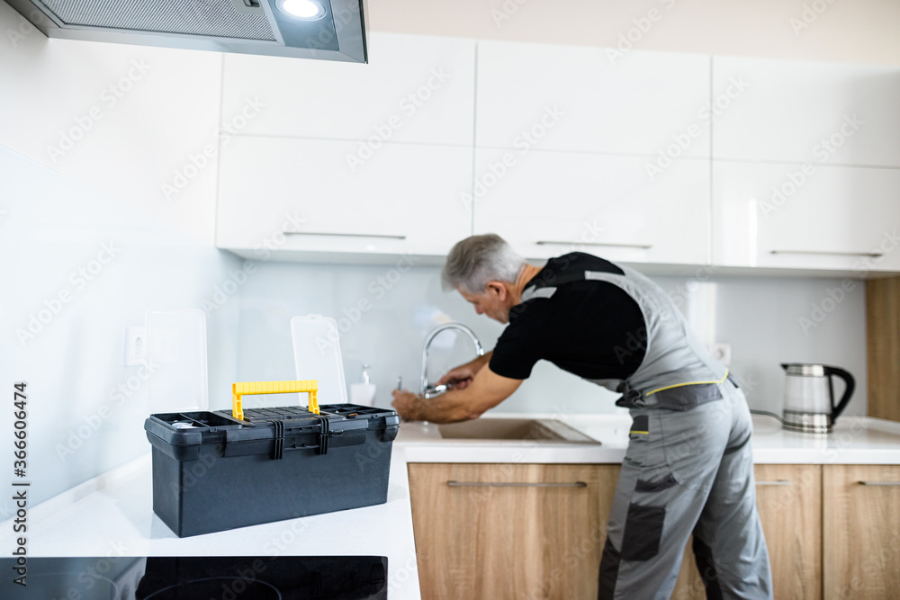 Aged repairman in uniform working, fixing broken kitchen tap using adjustable wrench. Repair service concept Aged repairman in uniform working, fixing broken kitchen tap using adjustable wrench. Repair service concept