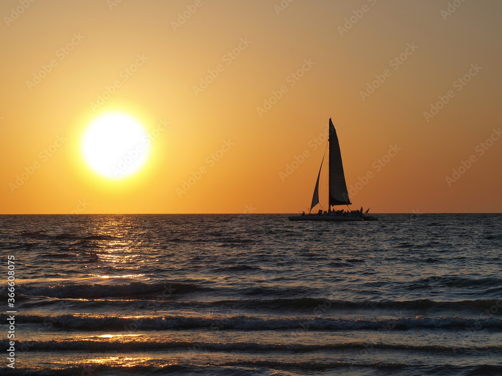 Sunset Sailing on the ocean with the bright orange-red sky, a large sun low on the horizon and a silhouette of a sailboat