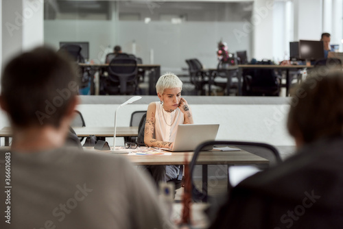 Working on a new project. Focused young and beautiful blonde tattooed businesswoman looking using laptop and thinking about something while sitting at her working place in modern office