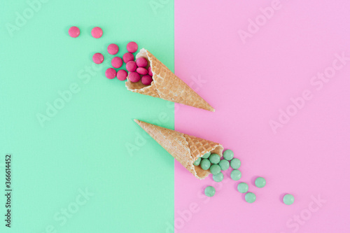 Two ice cream cones centre frame with candy sweets spilling out. Complementary colours of green and pink sweets and background. Food photo in flat lay style against vivid background with copy space