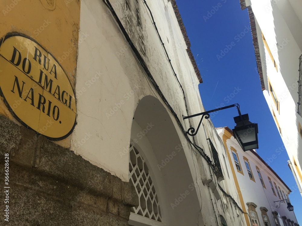 Fototapeta premium Surreal view of the ancient Imaginary Street -- Rua do Imaginario, Évora, Portugal. Ancient yellow and white architecture along narrow, crooked street against blue sky. High quality photo in natural