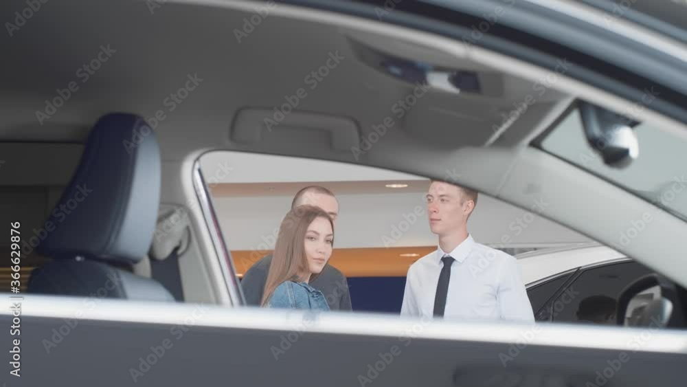 The selective focus of a friendly car dealer shows and explains to a young married couple all the pros and cons of a brand new car at the dealership The selective focus of a friendly car dealer shows and explains to a young married couple all the pros and cons of a brand new car at the dealership