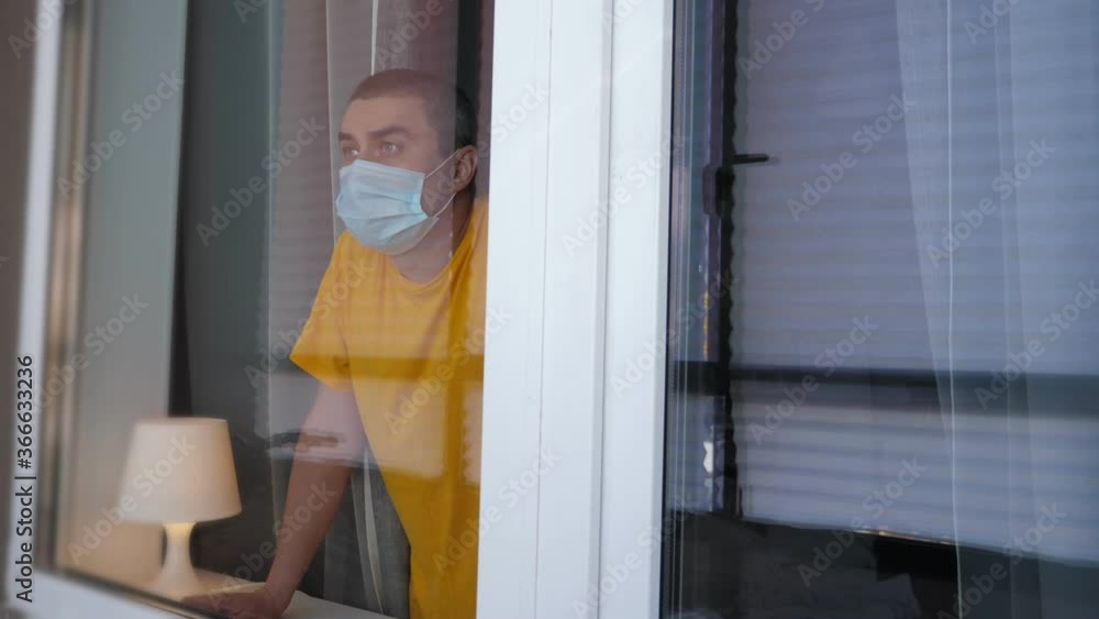 COVID-19 Pandemic Coronavirus. Quarantine man in medical mask on face looking through the window. People on self isolation concept COVID-19 Pandemic Coronavirus. Quarantine man in medical mask on face looking through the window. People on self isolation concept