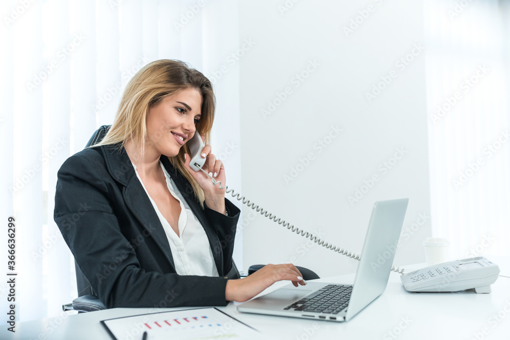 Smiling female entrepreneur with blonde hair talking on office phone and looking at laptop. Smiling female entrepreneur with blonde hair talking on office phone and looking at laptop.