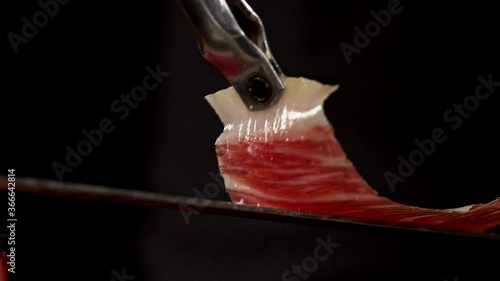 tongs grabs a slice of serrano Iberico ham from a plate,  in a short image detail. Very appetizing to eat that delicacy, with impressive red and white colors photo