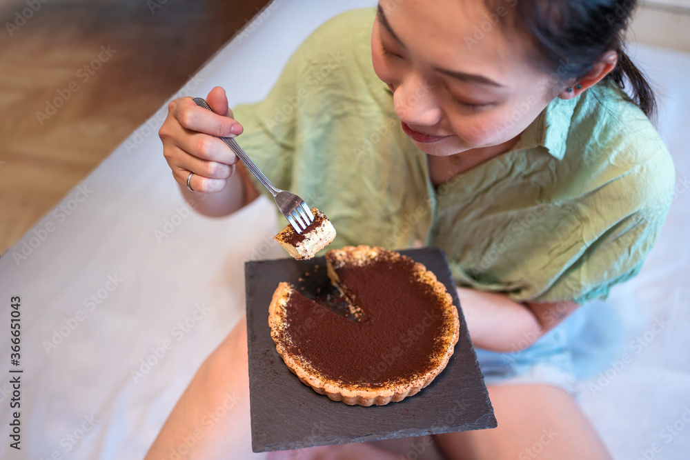 A top view of young pretty Asian woman eating delicious chocolate tart on bed