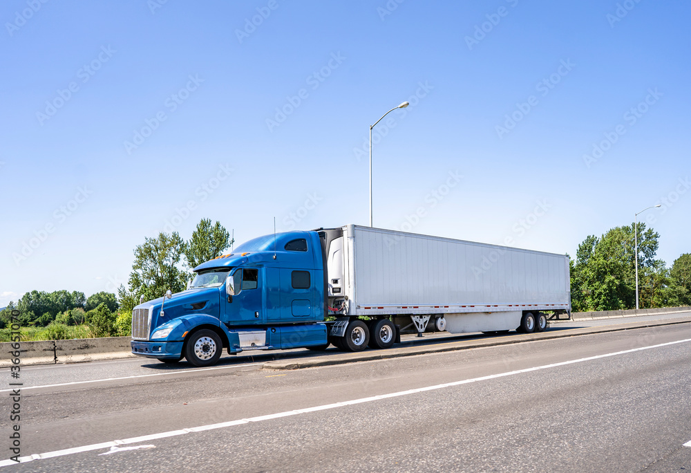 Blue big rig classic semi truck with high cab transporting load in refrigerator semi trailer running on the highway road entrance intersection Blue big rig classic semi truck with high cab transporting load in refrigerator semi trailer running on the highway road entrance intersection