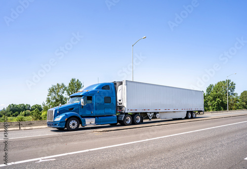 Blue big rig classic semi truck with high cab transporting load in refrigerator semi trailer running on the highway road entrance intersection