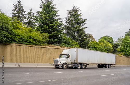 Big rig white bonnet day cab semi truck transporting commercial cargo in semi trailer moving on the highway road with concrete wall on the side