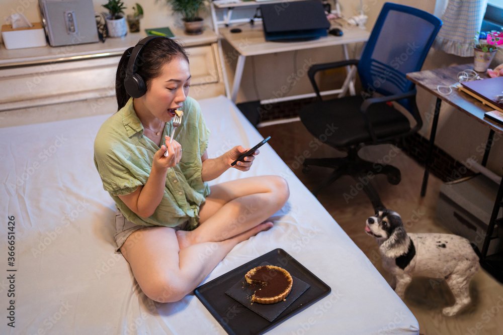 Young pretty Asian woman eating delicious chocolate tart on bed