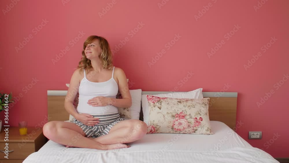 A smiling pregnant woman is sitting in bed with her hands around her belly. A smiling pregnant woman is sitting in bed with her hands around her belly.