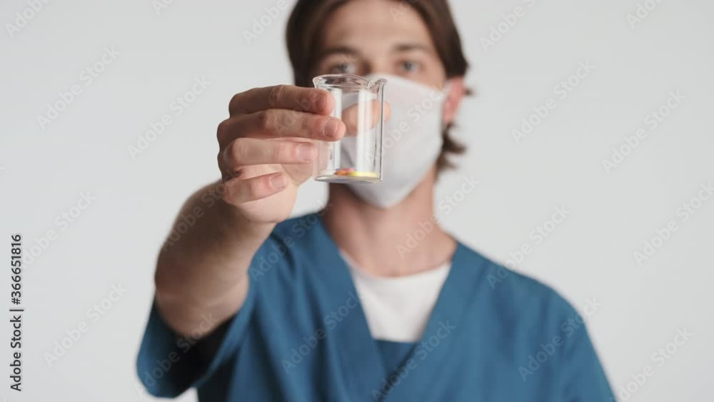 Young attractive male doctor in mask confidently showing medical pills on camera over white background. Safety concept Young attractive male doctor in mask confidently showing medical pills on camera over white background. Safety concept
