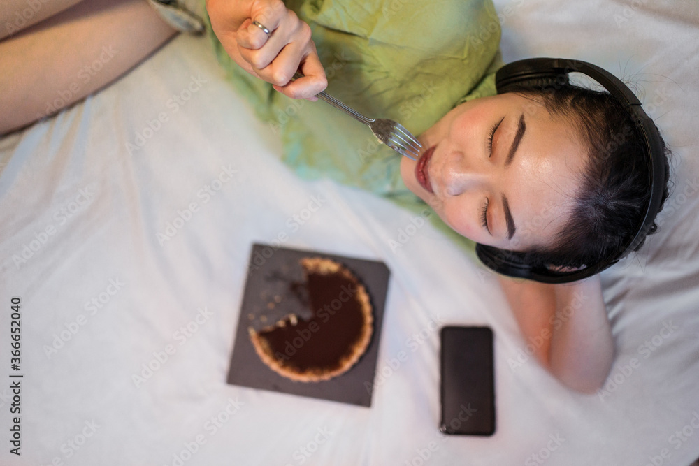A top view of young pretty Asian woman eating delicious chocolate tart on bed