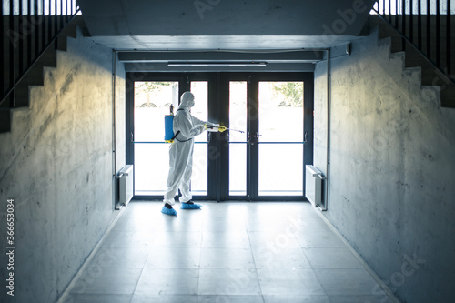 A man wearing protective disinfection suit and a spray stands in front of a glass doors under the staircases. Worker cleaning up the business center. Healthcare  covid-19 concept.