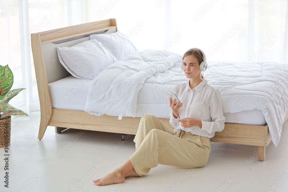 Young attractive caucasian woman wearing white casual clothes with headphone. She listening music or radio by using cell phone or music player in bedroom. (relaxation with technology concept)