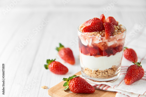 Homemade layered dessert with fresh strawberries, cream cheese or yogurt, granola and strawberry jam in glass on white wood background. Healthy organic breakfast or snack concept. Selective focus.