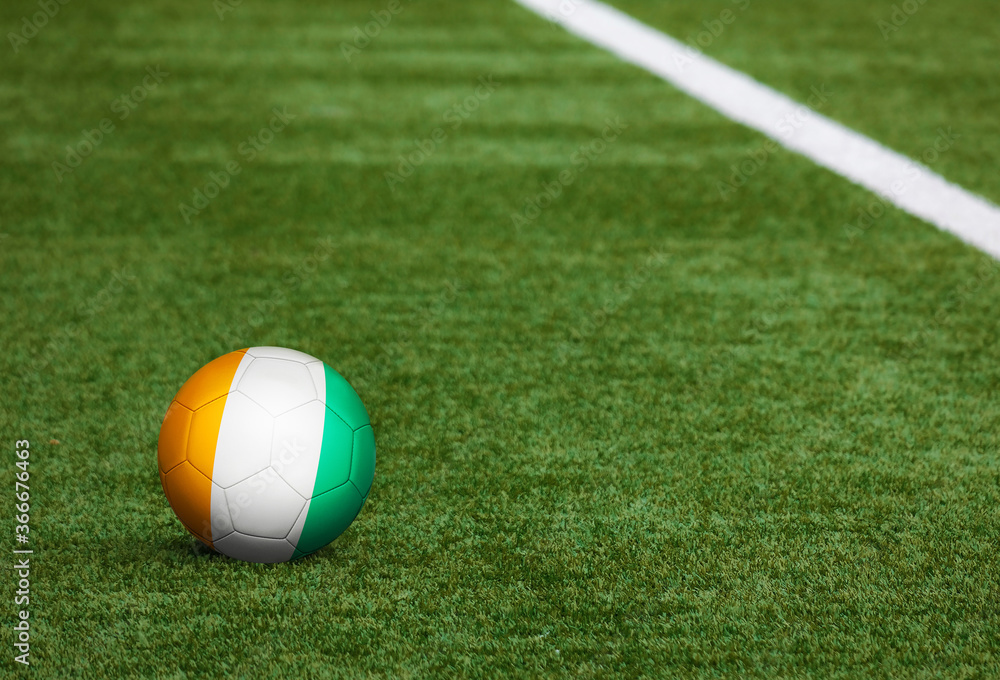 Naklejka Flaga Wybrzeża Kości Słoniowej na piłkę w tle boisko do piłki nożnej. Narodowy motyw piłki nożnej na zielonej trawie. Koncepcja zawodów sportowych.