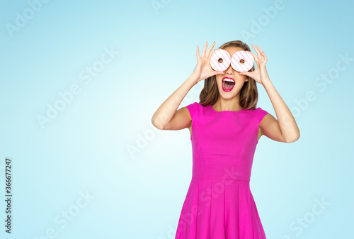 people and fast food concept - happy young woman or teen girl in pink dress having fun and looking through donuts over blue background