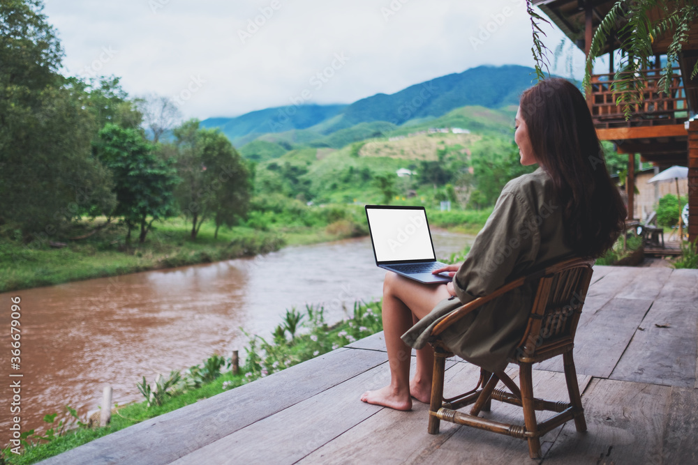 Mockup image of a young asian woman working on laptop computer while sitting by the river with mountains and nature background Mockup image of a young asian woman working on laptop computer while sitting by the river with mountains and nature background