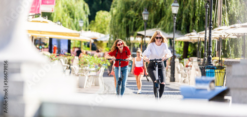 Trendy fashinable teenager girls riding public rental electric scooters in urban city environment. New eco-friendly modern public city transport in Ljubljana, Slovenia.