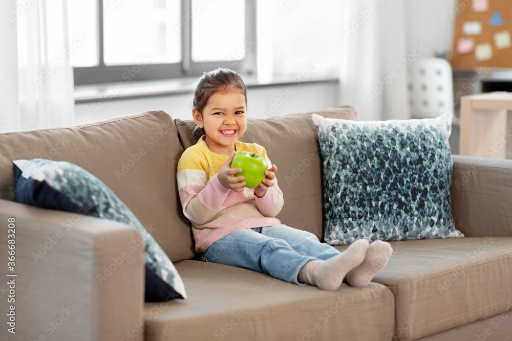 healthy eating and childhood concept - happy smiling little girl with green apple sitting on sofa at home healthy eating and childhood concept - happy smiling little girl with green apple sitting on sofa at home
