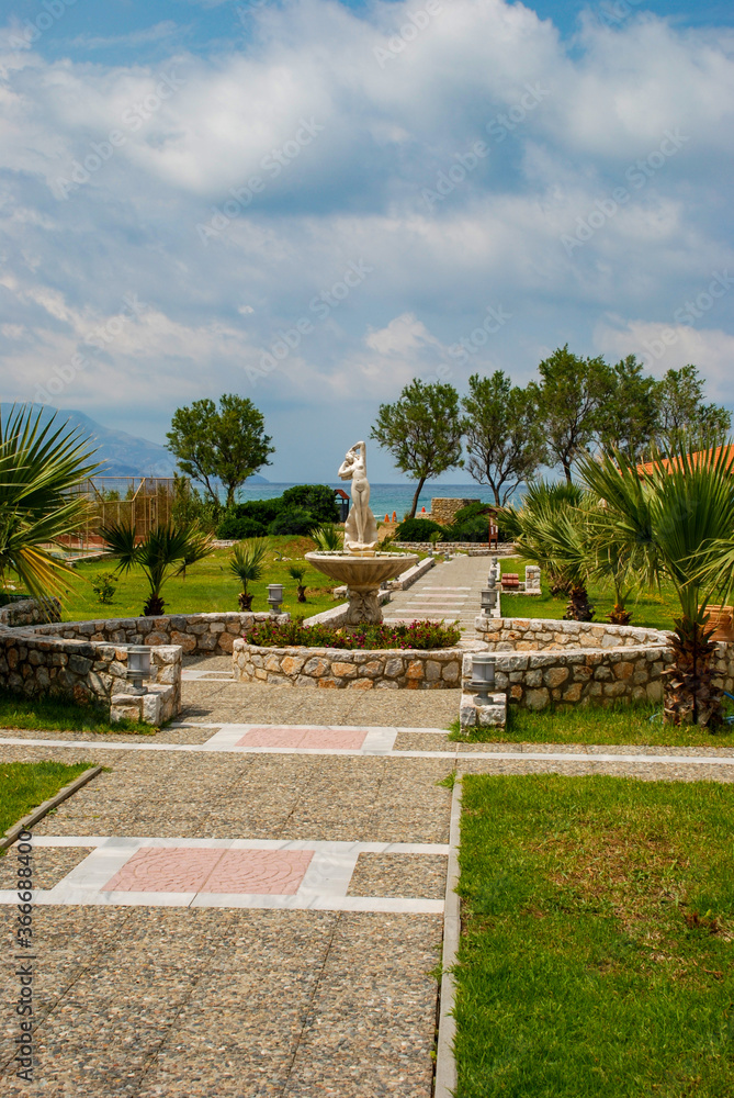 A park alley by the warm sea in summer in cloudy weather, geometric patterns on the path and a Greek statue in the center.