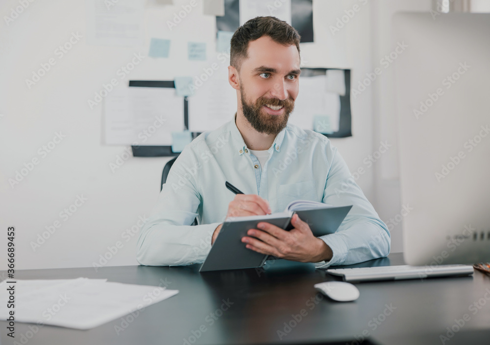 young bearded brunette smiling man looking happy while taking notes to his agenda working on business project in his modern office, work routine concept young bearded brunette smiling man looking happy while taking notes to his agenda working on business project in his modern office, work routine concept