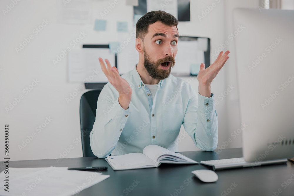 young bearded brunette man looking surprised while working on business project in his modern office, holds hands in the air, work routine concept