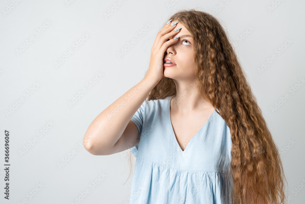 Photo of tired European young woman with curly long hair, covers face, rolled eyes, feels fatigue, needs good rest, has sleepy expression, isolated over white background. Weariness and people