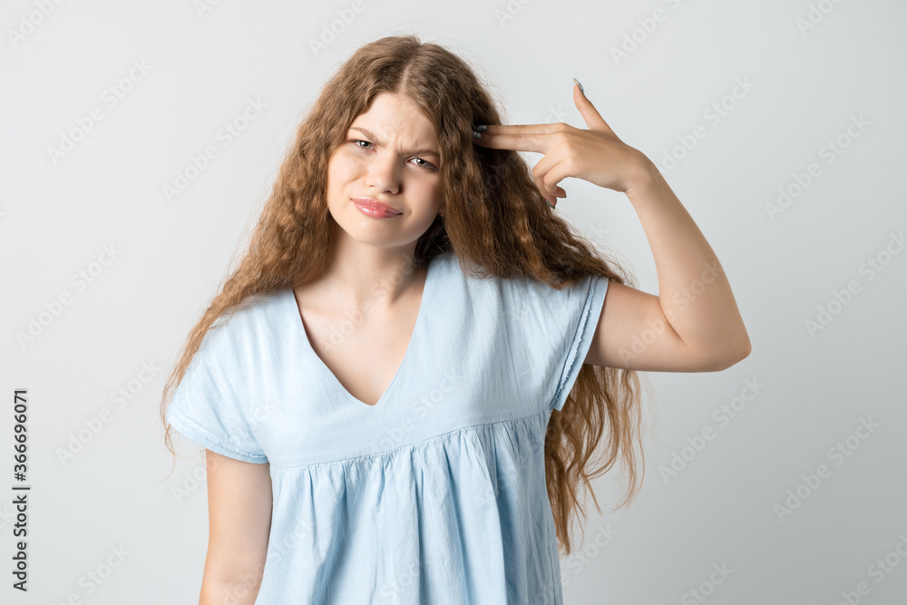 I'm fed up with this! Stylish European girl with curly long hair shoots in temple, tilts head, dressed in casual t shirt, demonstrates suicide gesture, isolated on white background I'm fed up with this! Stylish European girl with curly long hair shoots in temple, tilts head, dressed in casual t shirt, demonstrates suicide gesture, isolated on white background