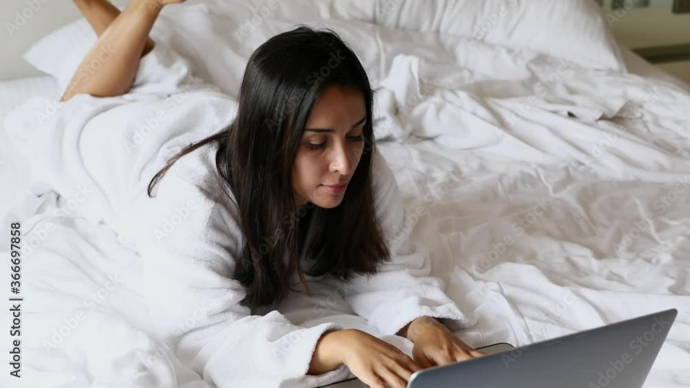 Smiling brunette woman wearing bathrobe in white morning bed, chatting, working, communicate online, looking at open laptop screen Smiling brunette woman wearing bathrobe in white morning bed, chatting, working, communicate online, looking at open laptop screen
