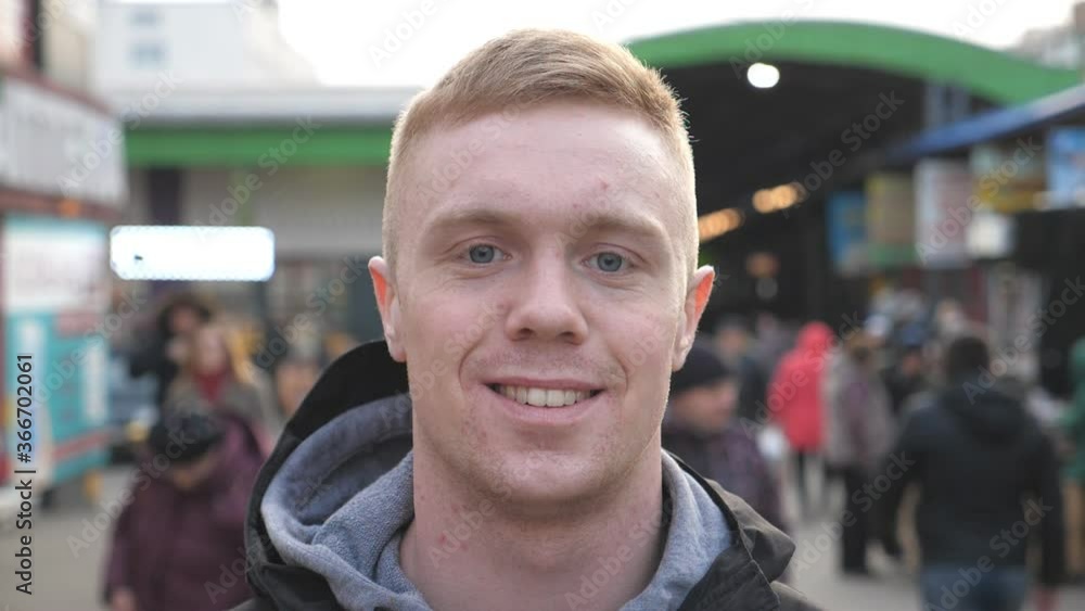 Portrait of young happy man standing at city street with positive emotions and feelings on his face. Guy looking and smiling into camera in the crowdy. Blurred people moving around. Close up Portrait of young happy man standing at city street with positive emotions and feelings on his face. Guy looking and smiling into camera in the crowdy. Blurred people moving around. Close up