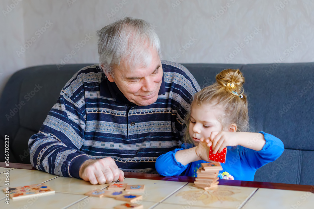 Naklejka premium Beautiful toddler girl and grandfather playing together pictures memory table cards game at home. Cute child and senior man having fun together. Happy family indoors