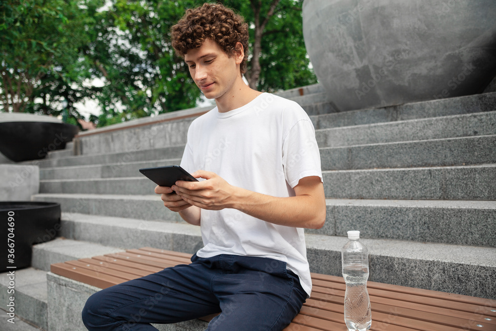 Obraz premium Portrait of a young curly male wearing white t-shirt in a city park, using digital tablet. Freelance work outdoor. Empty copy space.