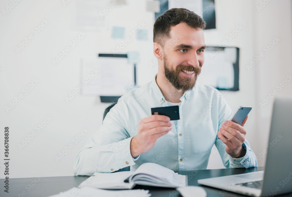 handsome smiling bearded businessman in elegant shirt sits in office showing bank card looking excited while working on business project , money making motivation concept