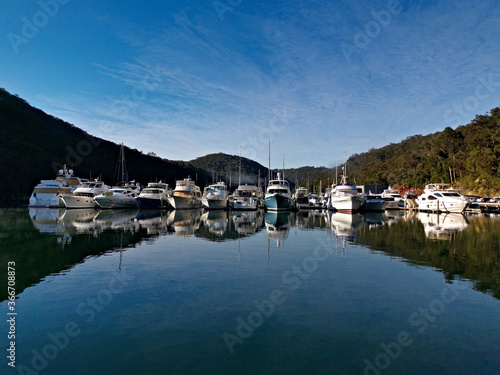 Early morning view of a creek with beautiful reflections of blue sky, luxury boats, mountains and trees on water, Cowan Creek, Bobbin Head, Ku-ring-gai Chase National Park, New South Wales Australia © Ivan