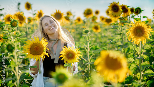 Beautiful young blonde woman in a hat and white shirt walks and laughs in a sunflower field