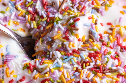 Close-up of a mug with yoghurt and colored sprinkles made of sugar, a spoon in the white cream, perfect child dessert for after lunch