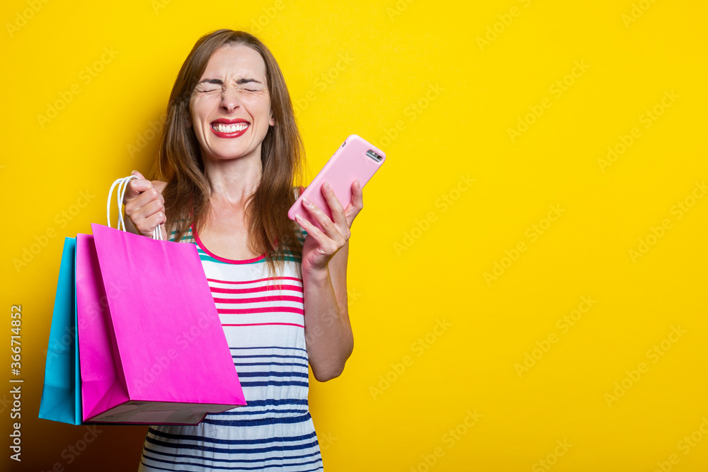 Joyful squinting young girl with phone holding shopping bags on yellow background. Joyful squinting young girl with phone holding shopping bags on yellow background.