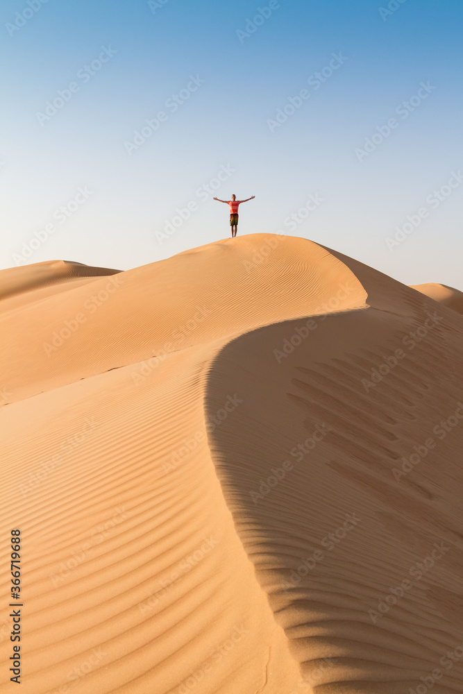 Male traveler standing on the top of dune, arms up to the sky, while traveling sand dessert in Oman. Travel adventure concept. - obrazy, fototapety, plakaty 