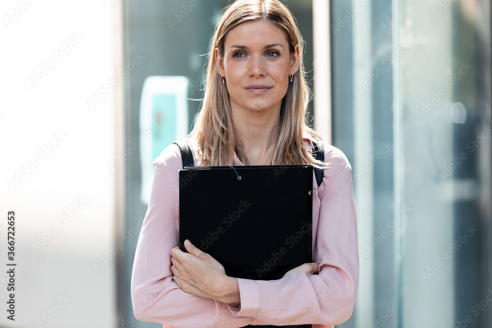 Fototapeta premium Beautiful blonde female student looking to side outdoors while holding a folder on campus of university.