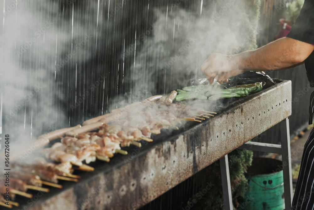 Hand of young man grilling some meat and vegetables on long rectangular barbecue with smoke on the street outdoor. Summer barbecue cooking. Barbecue with raw vegetables. BBQ on the terrace Hand of young man grilling some meat and vegetables on long rectangular barbecue with smoke on the street outdoor. Summer barbecue cooking. Barbecue with raw vegetables. BBQ on the terrace