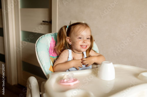 Little girl 2 years old with ponytail hairstyle plays with water, laughs and smiles, sitting in a highchair in the kitchen at home 