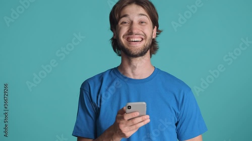 Attractive bearded guy using smartphone and joyfully looking in camera over colorful background