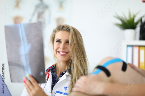 Woman doctor is holding an X-ray picture next to patient with kinesio tape on her knee. Injuries and sprains in athletes concept photo