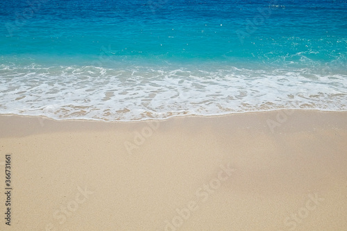 Soft ocean wave with foam licking the beautiful golden sandy beach shore. Close up of turquoise clear sea water texture on a windless and sunny summer day. Tropical background, copy space, top view.