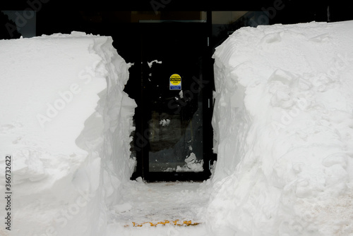 A path cut through a snowbank leading to a door. The sign on the door says CAUTION AUTOMATIC DOOR ACTIVATE SWITCH TO OPERATE. Interior of building behind door is dark.