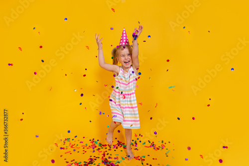 Happy smiling birthday child girl in pink cup surrounded by flying confetti jumping on colored yellow background . Celebration, childhood, emotions.