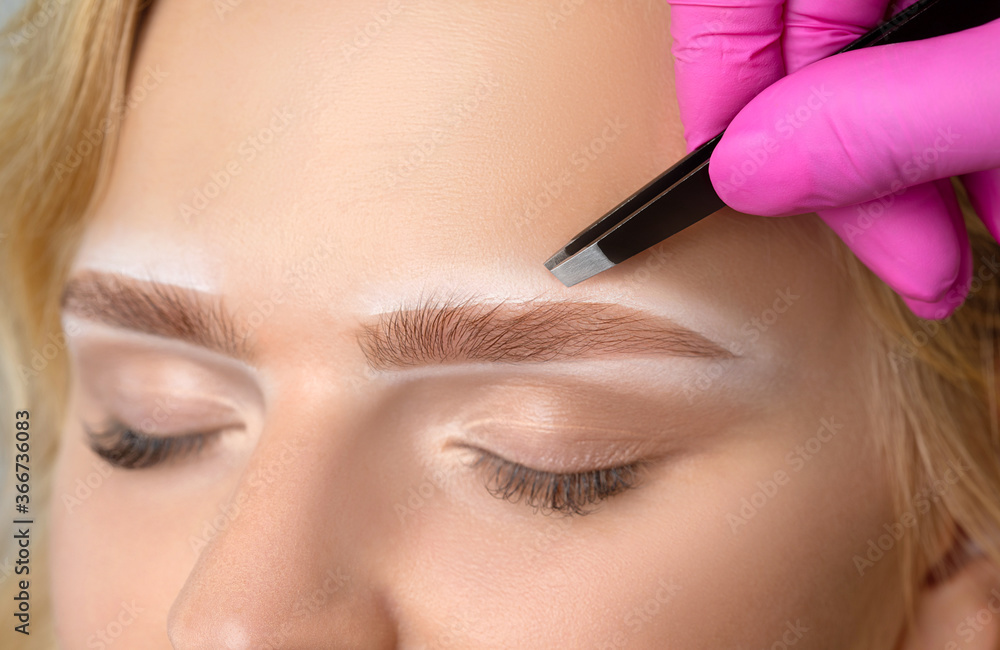 Beautiful blond woman with curly hair having Permanent Make-up Tattoo on her Eyebrows. Eyelash artist plucks eyebrows with tweezers. Professional makeup and cosmetology skin care.